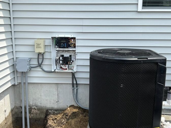Outdoor air conditioning unit next to a house with an electrical box mounted on the siding, cover open to expose wiring and circuit breakers.