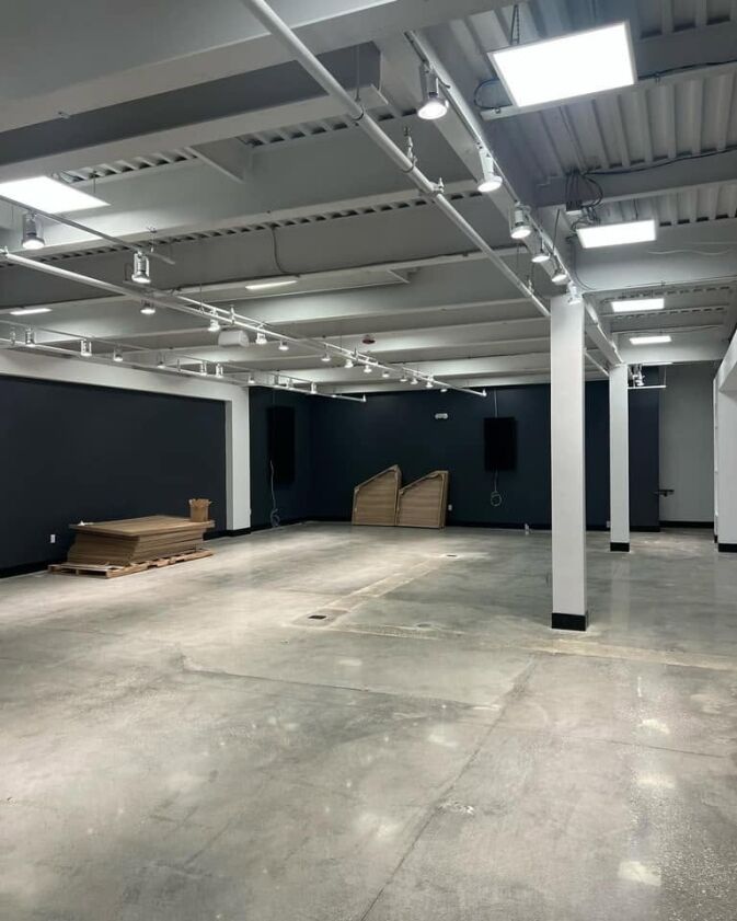 Empty industrial-style room with exposed ceiling, concrete floor, dark walls, overhead lighting, stacked wood, and cardboard boxes in one corner.
