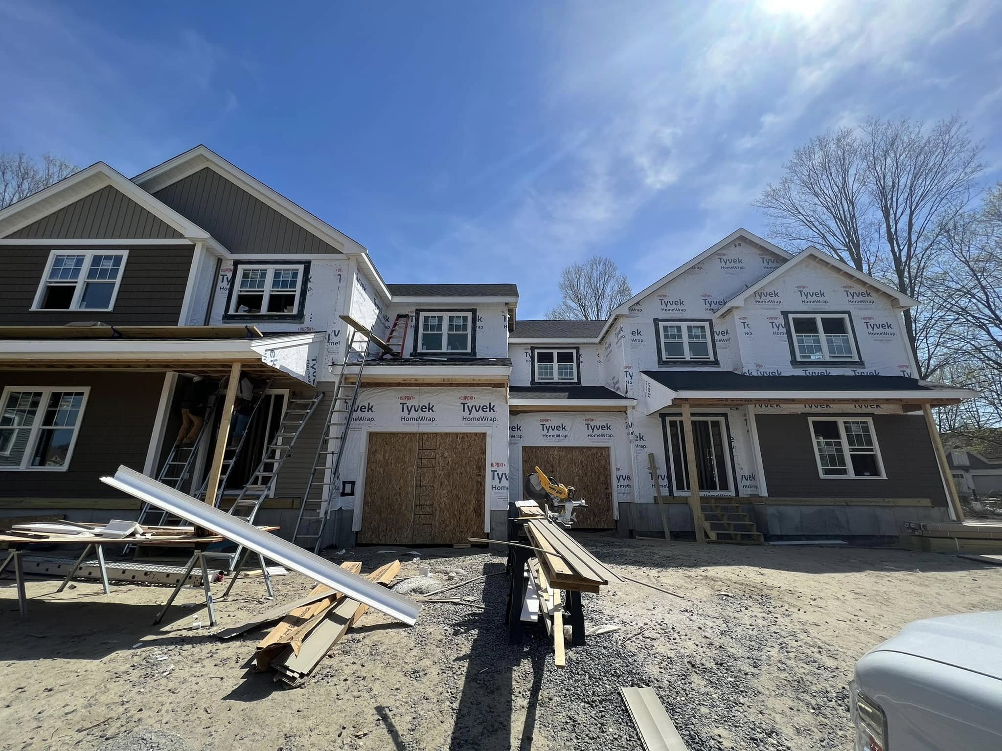 Two attached houses under construction, with some sections covered in Tyvek house wrap and building materials scattered in the foreground.