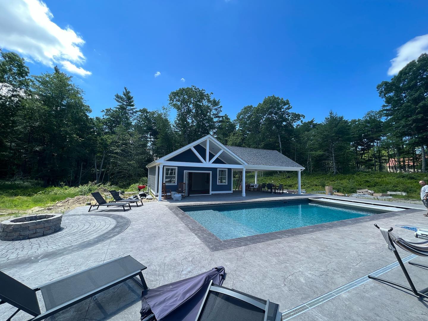A modern backyard with a pool, lounge chairs, fire pit, and a small pool house surrounded by trees under a bright blue sky.
